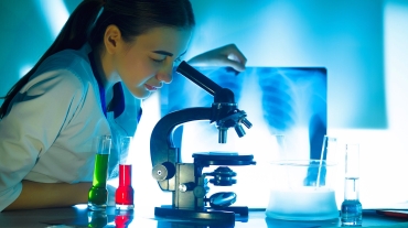Student girl looking in a microscope, science laboratory concept. Portrait of beautiful young woman in a laboratory sitting on her workplace.