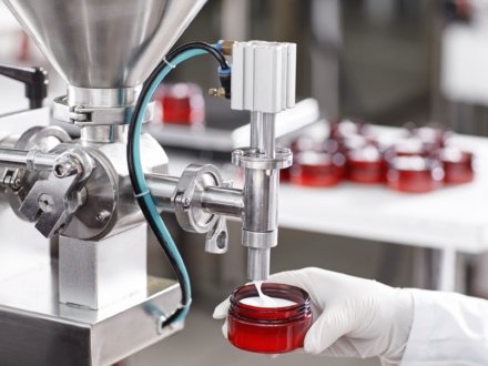 Hard mechanism of packing ready beauty cream into red container. A factory worker dressed in white lab gown and gloves holding red container under tap pouring ready cream. Production process of cream